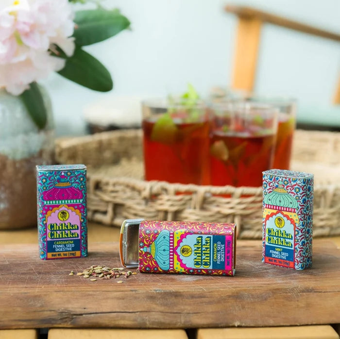 Three colorful tin containers of Chikka Chikka digestive snacks with their respective flavors indicated, placed on a wooden surface with a glass of a red beverage and a basket in the background.