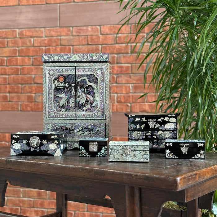 A group shot of various sized lacquer jewelry boxes with mother of pearl inlay sitting on a wood table next to a plant with a brick wall in the background