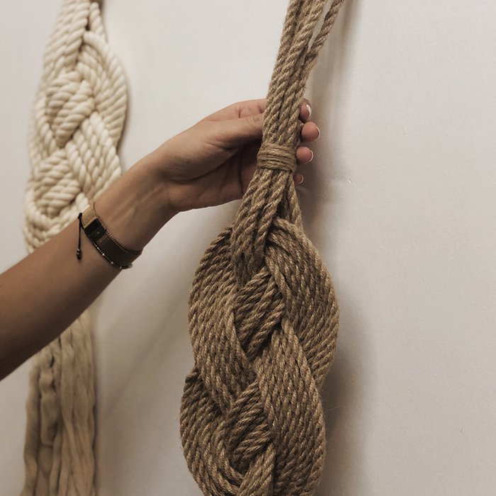 A hand holding a braided wall hanging made of natural jute fiber rope, with other similar hanging pieces in the background.