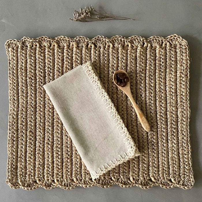 A rectangular, beige, handwoven placemat with a simple pattern. There is a folded cloth napkin and a carved wooden spoon on top.